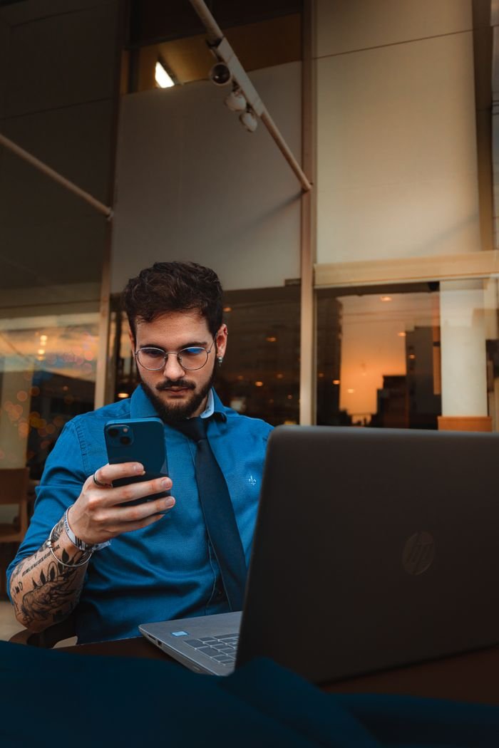 about-01 A man with a beard and eyeglasses focuses on his smartphone, sitting indoors with a laptop.